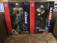 Front view of boxed Troy Aikman and Emmitt Smith McFarlane Toys figures, both new and sealed, sitting side by side on couch.