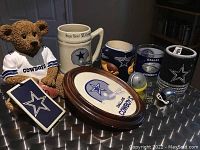 Full view of 10 Dallas Cowboys collectibles on table including teddy bear coin bank, mugs, plaque, mini helmets, koozie, pin button, and tag.