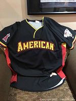 Front view of black American League All Star baseball jersey with team name 'AMERICAN' in large yellow and red letters, Angels logo patch on right sleeve, and All Star logo patch on left sleeve.