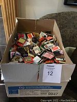 Box filled with a large quantity of assorted matchbooks piled inside a cardboard box.