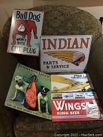 Overview photo showing four metal advertisement signs from Bull Dog Cut Plug, Indian Motorcycle, Tennents Lager, and Wings Cigarettes arranged on a couch.