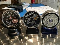 Three commemorative hockey pucks with printed game details and team logos, each displayed on stands on a metal surface.