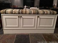 Front view of white wooden bench with three storage doors and striped cushion on top.