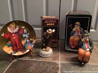 Photo showing three clown-themed items: a decorative plate, a small figurine of a clown looking down, and a larger seated clown figurine with a cane, all displayed on a floor.