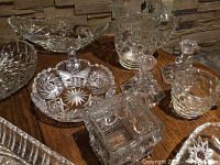 Photo of assorted crystal glassware including pedestal bowl, oval platter, covered dishes, candle holders, and pitcher on wooden table against stone wall background.