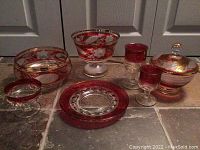 Seven pieces of red crystal glassware including bowls, compote, and plates with floral and gold detailing on tile floor