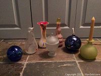 Photo showing seven assorted glass items including vases, spherical objects, and candle holders on a tile floor.