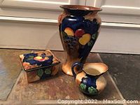 Three pieces of H & K Tunstall pottery displayed on tile floor: a large vase, small pitcher, and covered square box, all decorated with fruit and leaves in cobalt blue and beige glaze.