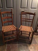Two hardwood chairs with woven seats placed side by side on a tiled floor, showing overall condition and ladder back design.