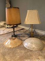 Two table lamps, one with a brown cylindrical shade and turned wooden base and another with a beige tapered rectangular shade and brass square base, both placed on carpeted floor with two clear etched glass lamp shades in front, one with yellow tint and one clear.