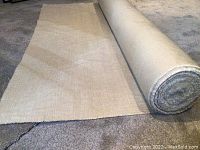 Wide view of beige fabric bolt partially unrolled on carpeted floor showing fabric texture and length.