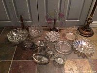 Lot of 11 assorted used crystal bowls and dishes with various shapes and patterns, shown on tile floor next to lamp base.