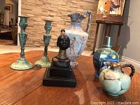 Front view of all 6 decorative items grouped together on table showing details: two green patina metal candlesticks, a tall bird motif pitcher, black bust sculpture, blue-green Goebel Lampe Berger, and small turquoise pitcher with brown trim.