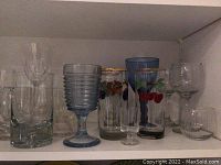 Shelf with variety of used clear and colored drinking glasses, goblets, and small bowls with some decorative painted designs.