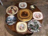 Eight decorative plates with various classical, scenic, and abstract designs arranged on a wooden table.