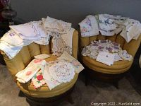 Wide view showing many folded embroidered and crocheted linens on two yellow chairs