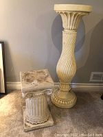 Photo of two decorative pillars/stands, a taller cream-colored one with spiral fluted design and a smaller white fluted classical column stand with a square top, placed indoor on carpet