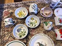 Overview of assorted porcelain bowls, mugs, and cups with floral and bird decoration laid on patterned carpet.