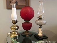 Three vintage style table lamps on glass table, showing red globe lamp, brass and white glass lamp, and another lamp with floral base.