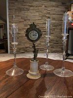 Full view of the vintage ornate clock and three clear crystal candle holders arranged on wooden table in front of brick wall background.