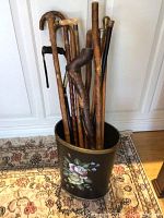 Multiple vintage wooden walking sticks placed standing inside a black cylindrical container with floral painting, showing wear and varying handle styles.