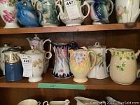 Shelf with eight coffee and cocoa pots, pitchers including varying floral patterns and decorative designs; two of the pieces have tags attached.