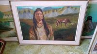 Framed painting showing Native American man in front of teepees and grazing horse, placed on carpeted floor.
