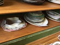 Multiple various plates and platters stacked on shelf showing different shapes and decorative patterns.