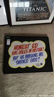 Frontal view of the framed Honest Ed poster on carpeted floor next to a Titanic poster.