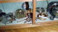 Photo showing a variety of silver plate items including biscuit barrel, candle holders, strainer, and serving plates inside a glass-front wooden cabinet.