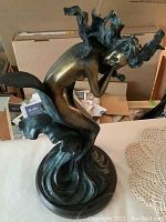 Side angle showing the mermaid statue with flowing hair seated on stylized waves and mounted on a round stone base.
