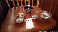 Four sterling silver items on a wooden table: a small round bowl with blue insert, two small bowls, and a tea strainer.