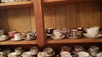 Shelf with multiple rows of assorted tea cups and saucers in floral and gold patterns.