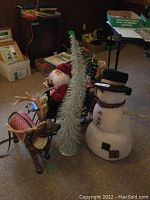 Photo showing the three Christmas decor items: stuffed snowman, silver tree, and decorative sleigh with reindeer and Santa.