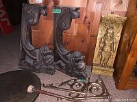 Photo of two large carved wood corbels with leaf designs, brass carved figure panel, the brass gong, and two cast aluminum wall brackets on floor against wood wall.