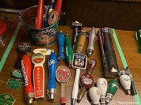 A variety of beer draught tap handles displaying multiple brand logos in different colors and materials placed on a table.