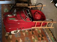 Side view of red metal ride-on fire truck pedal car showing white-wall wheels and one wood ladder