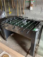 Foosball table viewed from an angle showing black metal sides, legs, and playing surface with players attached to rods.