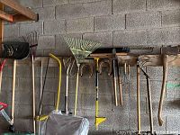 Wide view of assorted garden tools hung on a wooden plank attached to a cinder block wall, including rakes, hoes, shears, and horseshoes.