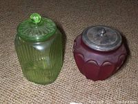 Two glass jars side by side on carpet: green ribbed glass jar with glass lid and red glass jar with flower pattern and metal lid.