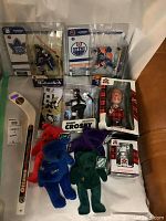Photo showing five sealed hockey figurines including Frank Mahovlich, Sidney Crosby, Edmonton Oilers player, two boxed figurines (lumberjack character and miniature hockey player), four plush collectible bears in red, blue, green, and purple, and a hockey mini stick marked Ottawa with #89.