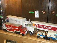 Photo showing a row of six toy tractor-trailers of varying sizes, including Coca-Cola and Maple Leaf brands prominently visible, providing scale and condition reference.