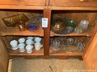 Photo of wooden shelf containing various vintage glassware and mugs including amber glass covered dish, clear glass bowls, and white mugs with designs.