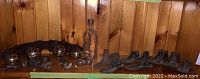 Photo of a shelf showing a vintage glass bottle, brass buttons, brass horse bells, and cast iron shoe forms arranged on a wooden shelf against a wooden wall.