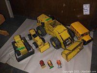 Photo showing all 7 vintage toy trucks arranged on paper. Includes large Mighty Loader, three medium construction vehicles, a yellow toy school bus, and three small miniature trucks.