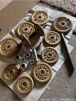 Photo showing multiple gold-colored cast iron weight plates branded 'WEIDER BARBELL' along with dumbbell handles and a barbell bar, laid out on a white sheet on the floor.