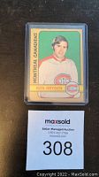 Photo of 1973 O-Pee-Chee NHL hockey card featuring Ken Dryden in protective sleeve with MaxSold lot number card visible.