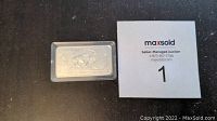 Top view of the silver-colored bullion bar in a plastic case next to a MaxSold auction identification card.