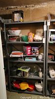 Photo of one metal utility shelf unit with five levels, filled with kitchenware and household items, against a wall.