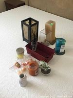Photo showing all items arranged on a bedspread: candles, candle holders, red woven basket, glass vase with stones, diffuser bottle, small plant, and accessories.